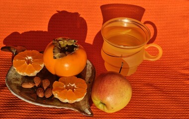 still life of fruits: persimmons and tangerine cut in half and almonds on a clay saucer. An orange cup and an apple on an orange napkin  background, healthy food concept