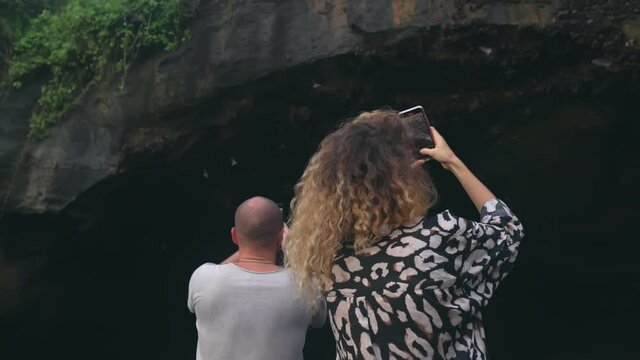 A girl and a man are shooting videos on their phones with a huge rocky cave from which bats fly out. Daytime slow motion. A couple of travelers take pictures of flying bats.