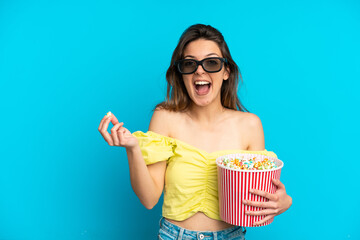 Obraz premium Young caucasian woman isolated on blue background with 3d glasses and holding a big bucket of popcorns