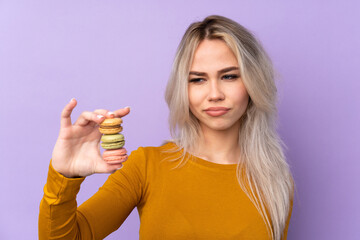 Teenager girl over isolated purple background holding colorful French macarons with sad expression
