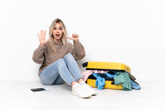 Teenager blonde girl with a suitcase full of clothes sitting on the floor counting six with fingers