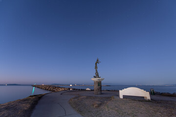 Fototapeta premium Twinkling stars in the dark morning sky shine over beacon statue positioned on the Ventura harbor mouth of the ocean