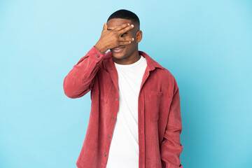 Young latin man isolated on blue background covering eyes by hands and smiling