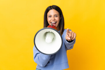 Caucasian girl isolated on yellow background shouting through a megaphone to announce something while pointing to the front
