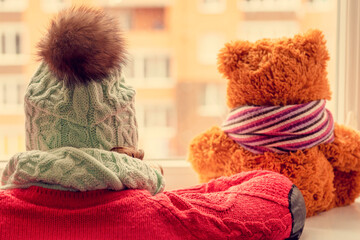 The girl in a cap at a window with  teddy bear. The woman in a warm cap and a toy bear cub look out of the window.