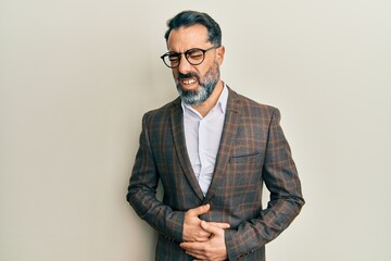 Middle age man with beard and grey hair wearing business jacket and glasses with hand on stomach because nausea, painful disease feeling unwell. ache concept. - obrazy, fototapety, plakaty