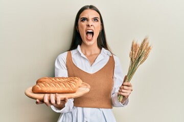 Beautiful brunette young woman holding homemade bread and spike wheat angry and mad screaming...