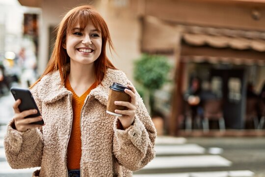 Young redhead girl smiling using smartphone drinking coffee at the city.