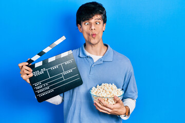 Handsome hipster young man eating popcorn holding cinema clapboard making fish face with mouth and...