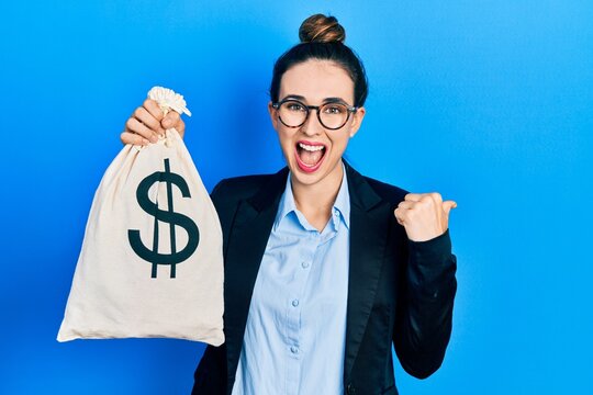 Young hispanic girl wearing business clothes holding dollars bag pointing thumb up to the side smiling happy with open mouth