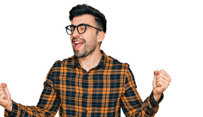 Hispanic man with beard wearing casual clothes and glasses very happy and excited doing winner gesture with arms raised, smiling and screaming for success. celebration concept.