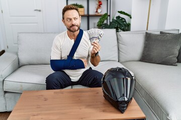 Middle age man with beard wearing arm on sling for motorbike accident holding insurance money...