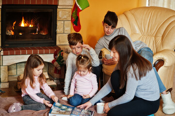 Happy young large family by a fireplace in warm living room on winter day. Mother with four kids at home read book.