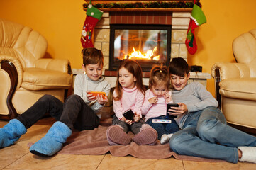 Happy four kids watching in they mobile phone at home by a fireplace in warm living room on winter day.