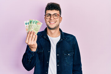 Young hispanic man holding argentine pesos banknotes looking positive and happy standing and smiling with a confident smile showing teeth