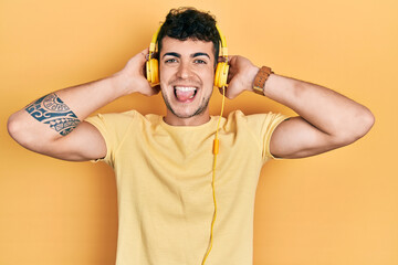 Fototapeta na wymiar Young hispanic man listening to music using headphones sticking tongue out happy with funny expression.