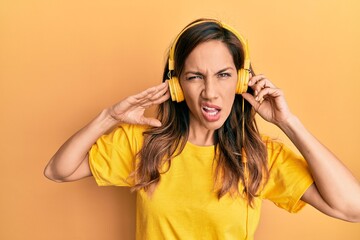 Young latin woman listening to music using headphones in shock face, looking skeptical and sarcastic, surprised with open mouth
