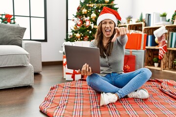 Young latin woman using laptop sitting by christmas tree pointing displeased and frustrated to the camera, angry and furious with you