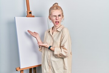 Beautiful caucasian woman with blond hair standing by painter easel stand presenting canva winking...