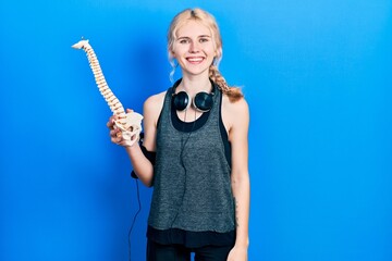 Beautiful caucasian sports woman with blond hair holding anatomical model of spinal column looking positive and happy standing and smiling with a confident smile showing teeth
