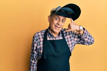Handsome senior man with grey hair wearing welding protection mask smiling doing phone gesture with hand and fingers like talking on the telephone. communicating concepts.