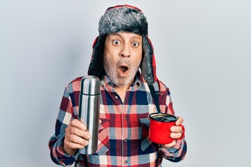 Handsome mature handyman wearing winter hat with ear flaps drinking hot coffee from thermo afraid...