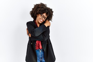 African american woman with afro hair wearing business jacket and glasses hugging oneself happy and...