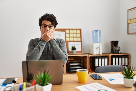 Young hispanic man wearing business style sitting on desk at office shocked covering mouth with hands for mistake. secret concept.