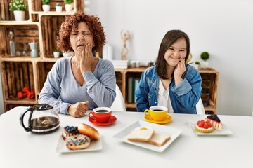 Family of mother and down syndrome daughter sitting at home eating breakfast touching mouth with hand with painful expression because of toothache or dental illness on teeth. dentist