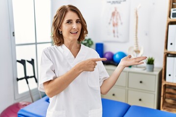 Fototapeta premium Middle age physiotherapist woman working at pain recovery clinic amazed and smiling to the camera while presenting with hand and pointing with finger.