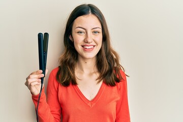 Young brunette woman holding hair straightener looking positive and happy standing and smiling with a confident smile showing teeth