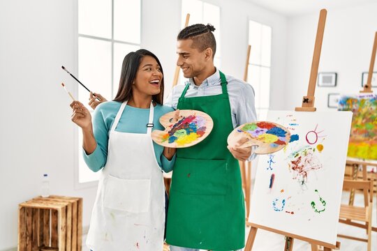 Young latin painter couple smiling happy holding paintbrush and palette at art studio