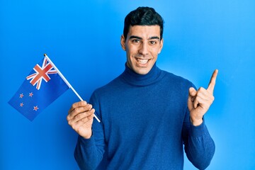Handsome hispanic man holding new zealand flag smiling happy pointing with hand and finger to the side