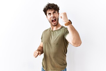 Hispanic man standing over isolated white background angry and mad raising fist frustrated and...