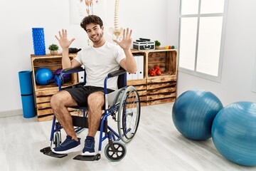 Hispanic man sitting on wheelchair at physiotherapy clinic showing and pointing up with fingers...
