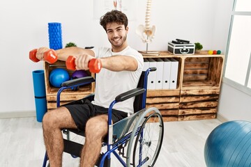 Young hispanic man sitting on wheelchair doing rehab using dumbbells at clinic