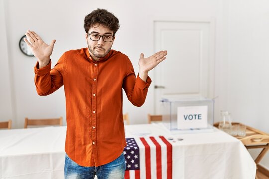 Hispanic Man Standing By Election Room Clueless And Confused Expression With Arms And Hands Raised. Doubt Concept.