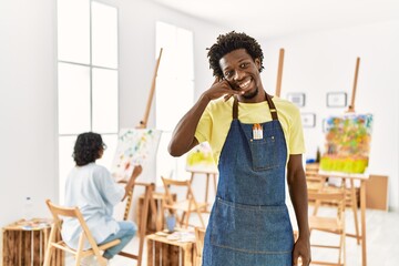 African young man standing at art studio smiling doing phone gesture with hand and fingers like talking on the telephone. communicating concepts.