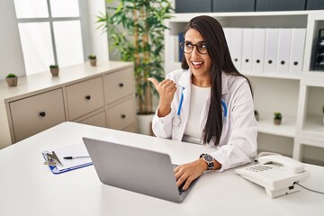 Young hispanic doctor woman doing video call at the clinic pointing thumb up to the side smiling happy with open mouth