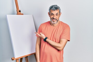 Handsome middle age man with grey hair standing by painter easel stand pointing aside worried and...