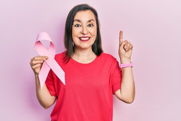 Middle age hispanic woman holding pink cancer ribbon surprised with an idea or question pointing finger with happy face, number one