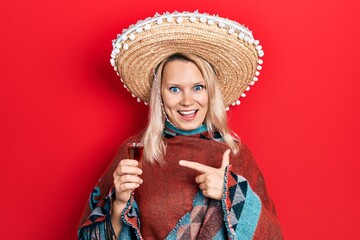 Beautiful caucasian blonde woman wearing festive mexican poncho drinking tequila shot smiling happy...