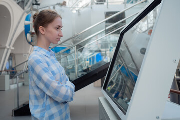 Portrait of woman in blue plaid shirt looking at interactive touchscreen display of electronic kiosk at futuristic exhibition or museum - side view. Education, technology concept