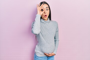 Beautiful woman with blue eyes expecting a baby, touching pregnant belly doing ok gesture shocked with surprised face, eye looking through fingers. unbelieving expression.