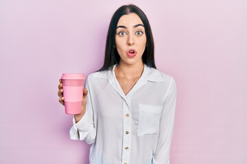 Beautiful woman with blue eyes drinking a take away cup of coffee scared and amazed with open mouth...