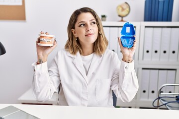 Young hispanic woman working at dentist clinic holding brackets and aligner smiling looking to the...