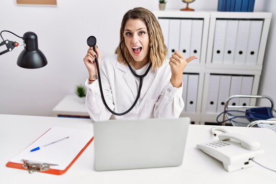Young hispanic woman wearing doctor uniform holding stethoscope at medical clinic pointing thumb up to the side smiling happy with open mouth