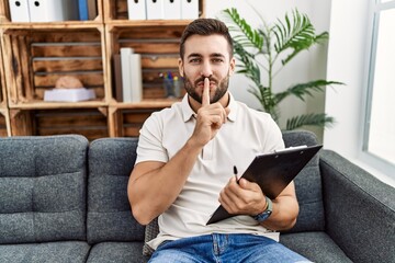 Handsome hispanic man holding clipboard working at psychology clinic asking to be quiet with finger...