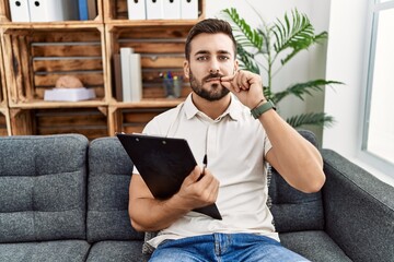 Handsome hispanic man holding clipboard working at psychology clinic mouth and lips shut as zip with fingers. secret and silent, taboo talking