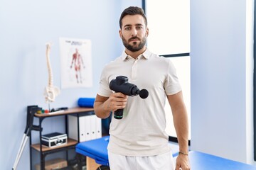 Naklejka na ściany i meble Handsome hispanic man holding therapy massage gun at physiotherapy center thinking attitude and sober expression looking self confident